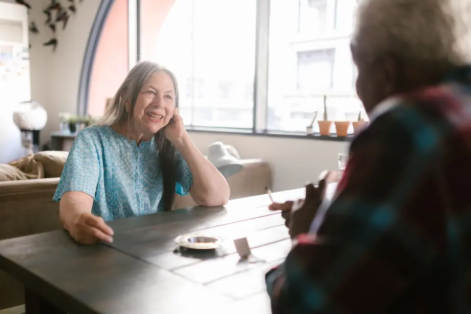 Een oudere vrouw praat glimlachend aan de keukentafel met een bezoeker