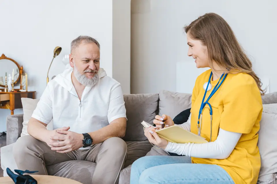 Een zorgverlener maakt aantekeningen tijdens een gesprek met een oudere man op de bank bij hem thuis